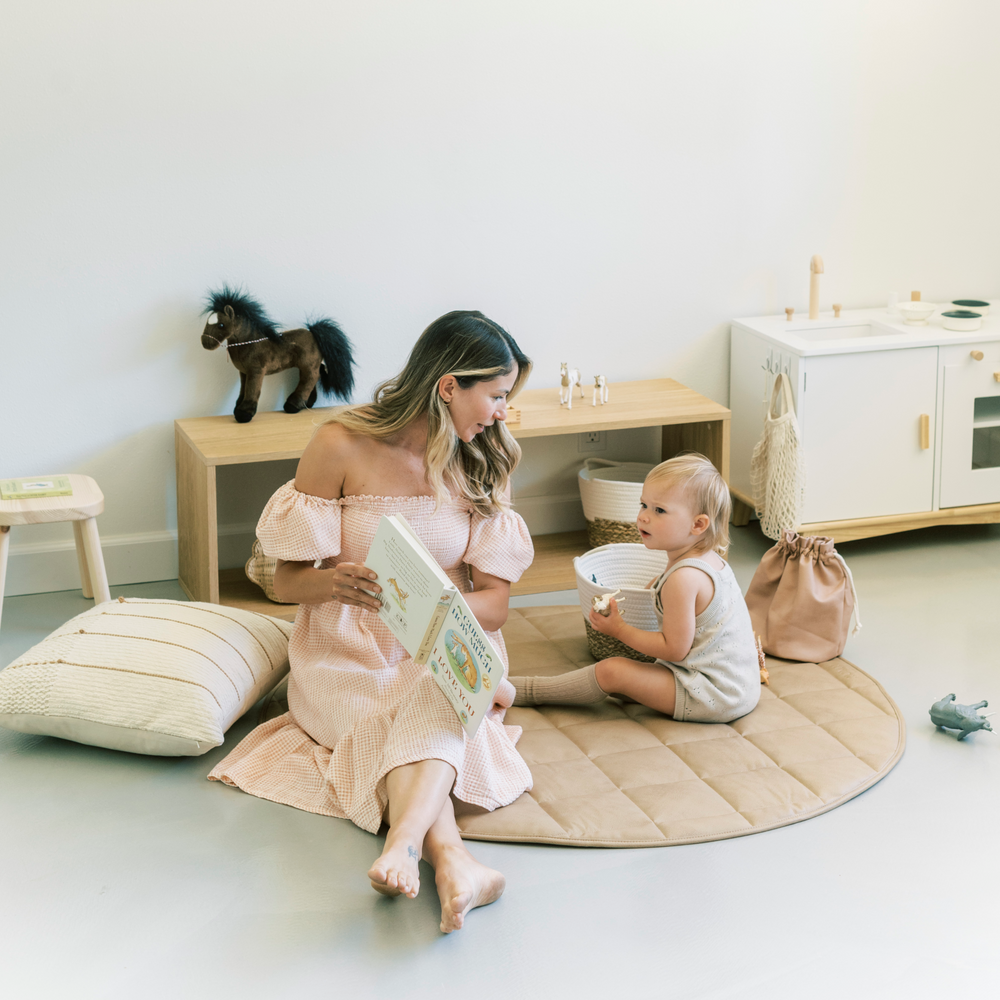 Photo of a mother sitting on a vegan leather play mat with her toddler daughter reading her a book.