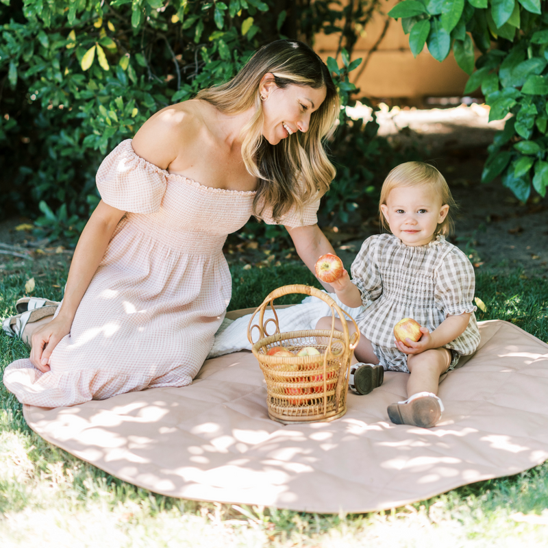 Outdoor photo of a smiling mother sitting on a vegan leather play mat with her smiling toddler daughter who is holding apples.