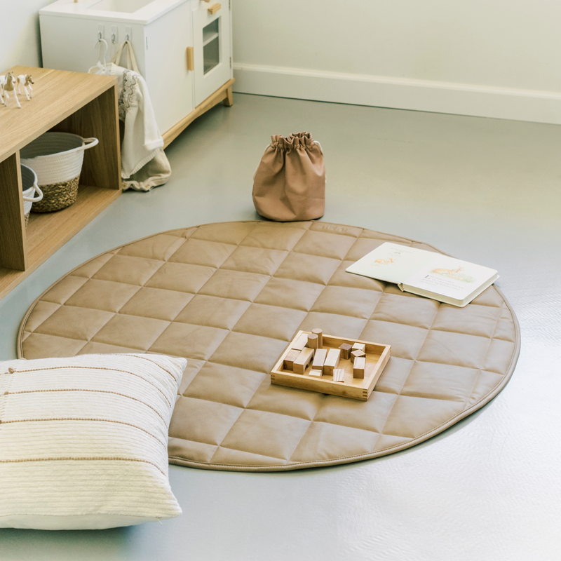 indoor photo of a child's play room with a vegan leather play mat on the floor with a book and wooden toys on it.