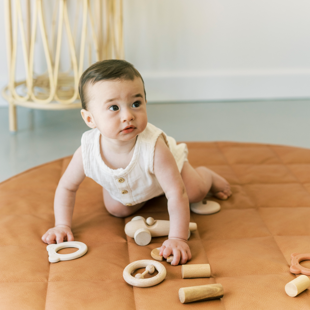 indoor photo of a baby with dark hair crawling on a vegan leather play mat amongst wooden toys.